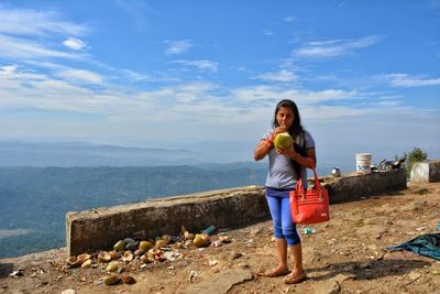 Portrait of young woman drinking coconut water while standing on mountain against sky