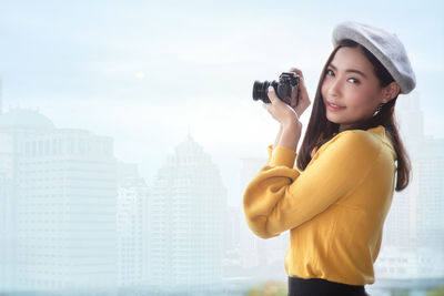 Side view of young woman photographing against modern city