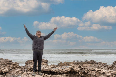Adult man with gray hair and glasses raising his arms to the sky above the rocks and the sea behind.