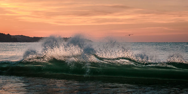 Sea waves splashing on shore against sky during sunset