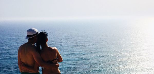 Rear view of shirtless man embracing girlfriend while looking at sea