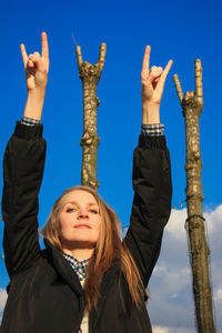 Portrait of woman showing rock sign with arms raised against blue sky