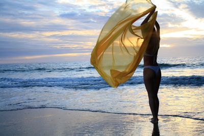 Young woman wearing bikini while holding yellow textile on shore at beach against sky during sunset