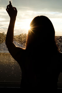 Rear view of silhouette woman standing by wet glass window during sunset