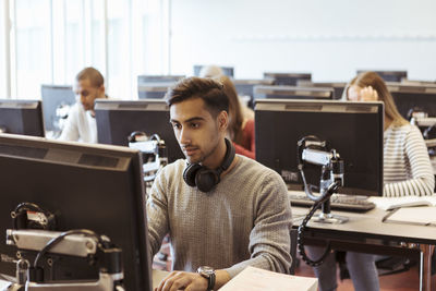 Serious young man using computer at desk with students in background at library