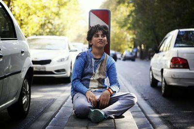 Full length of homeless boy looking away while sitting amidst street