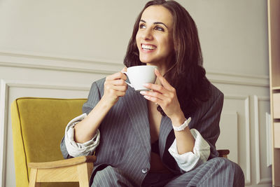 Ballerina in a gray striped suit with a cup of coffee smiling and dreaming is sitting in an armchair