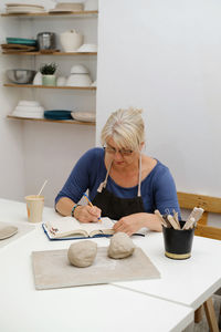 Woman potter planning work at workshop, writing in notebook in light studio her creations. clay