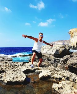 Happy man with arms outstretched standing on cliff by sea against sky