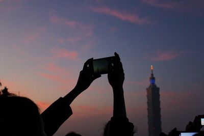 Cropped image of woman photographing taipei 101 against sky during sunset