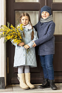 A couple of a boy and a girl of schoolchildren in spring coats with a bouquet of mimosa in  hands 