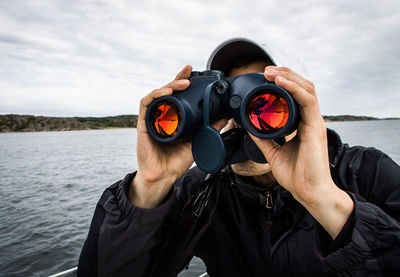 Man looking through binoculars in sea against cloudy sky