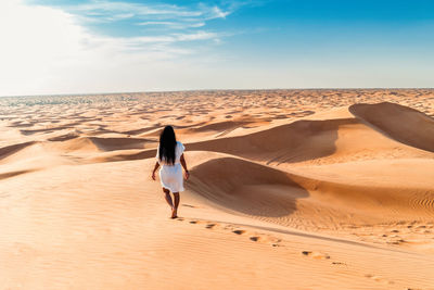 Rear view of woman walking on sand at beach against sky during sunset