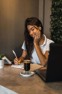 Cheerful hispanic female with earphones studying online and watching video lecture on laptop and taking notes in copybook at table with cup of coffee in cafe
