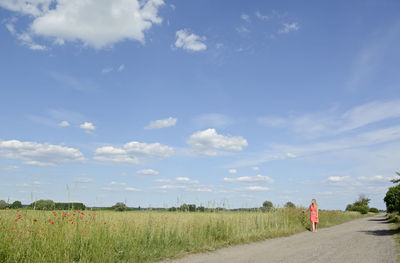 Woman walking on road by grassy field against sky