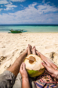 Low section of couple with coconut water relaxing at beach on sunny day