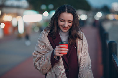 Happy young woman drinking drink with city in background at night