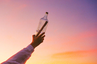 Cropped hand of woman holding bottle against sky during sunset