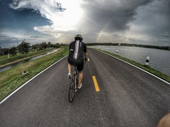 Rear view of man cycling on road by river against cloudy sky