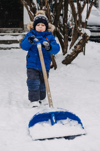 Little boy with a shovel in hand remove snow in backyard, snow removal. kid in blue jacket cleans