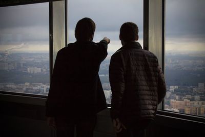 Rear view of man pointing at cityscape while friend looking through window against sky