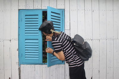 Side view of backpack woman peeking through blue window amidst wall