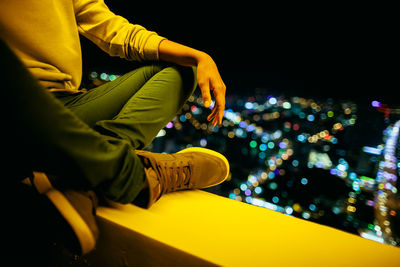 Low section of man sitting at balcony against illuminated city at night