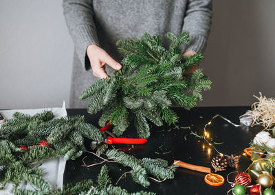 Young woman in warm grey knitted dress making diy fir christmas wreath in hand on table at home