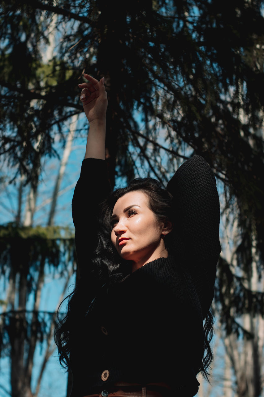 tree, one person, women, adult, young adult, plant, nature, spring, long hair, portrait, hairstyle, arms raised, clothing, forest, arm, lifestyles, leisure activity, outdoors, waist up, standing, person, female, sunlight, land, limb, photo shoot, blue, emotion, day, winter, looking, fashion, looking up, branch, human limb, brown hair, happiness