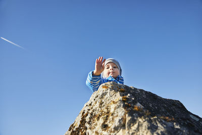 Cute boy on rock waving hand against sky