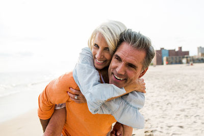 Happy man piggybacking woman at beach against clear sky