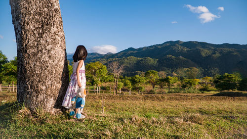 Girl holding toy while standing on grass by tree trunk against mountain and sky