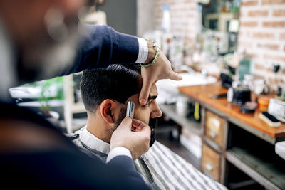 Crop anonymous barber using sharp straight blade for cutting hair of male customer in professional barbershop