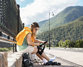 Young woman in yellow hoodie putting on protective equipment before roller skating on the embankment 