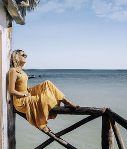 Woman sitting on railing by sea against sky