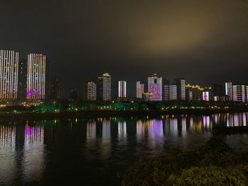 Illuminated buildings by river against sky at night