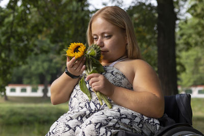 Smiling happy young woman with short stature on wheelchair holds, sniffing flowers in green park at