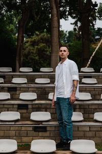 Portrait of young man standing against trees and outdoor seats