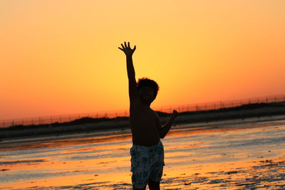 Boy with arms raised standing at beach during sunset