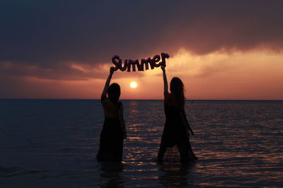 Silhouette women holding text while standing at shore against sunset sky