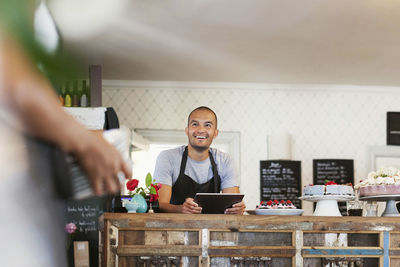 Smiling baker holding digital tablet while looking at female colleague in cafe