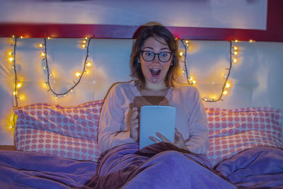 Young woman with mouth open using digital tablet while sitting on bed at home