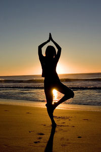 Silhouette woman doing tree pose yoga at beach against sky during sunset