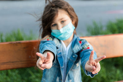 Caucasian girl sitting on a bench in the mask on the face with sad eyes and shows hands gesture viva