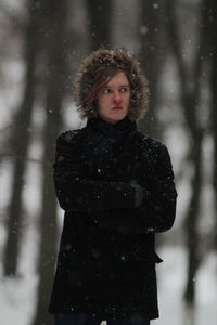 Thoughtful man with arms crossed standing amidst trees during winter