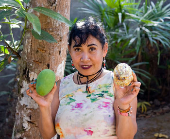 Portrait of woman holding fruits while standing by tree trunk