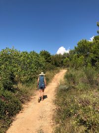 Rear view of mid adult woman wearing hat while walking on dirt road against blue sky during sunny day