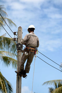 Rear view of man working on electricity pylon against sky