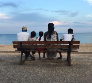 Full length rear view of family sitting on bench at beach against sky