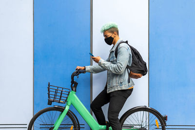 Side view of ethnic male hipster wearing face mask with blue hair sitting on bicycle and browsing cellphone in city near vivid orange build during coronavirus pandemic